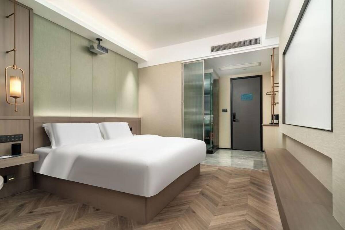 A modern hotel room with a large bed, a bedside table, a wall-mounted light, and a door leading to another room.