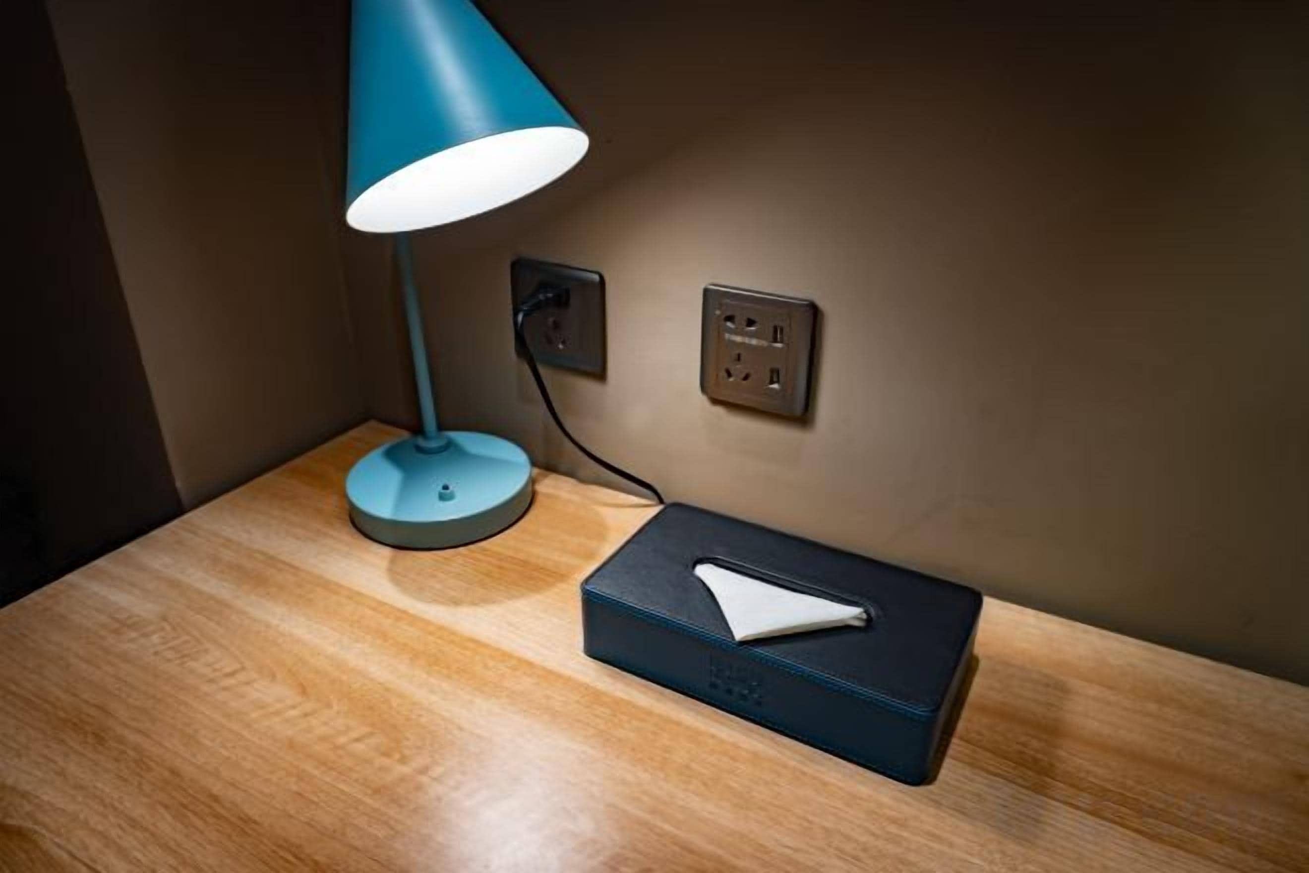 A wooden bedside table with a blue desk lamp, a power outlet, and a black box with a white object on it.