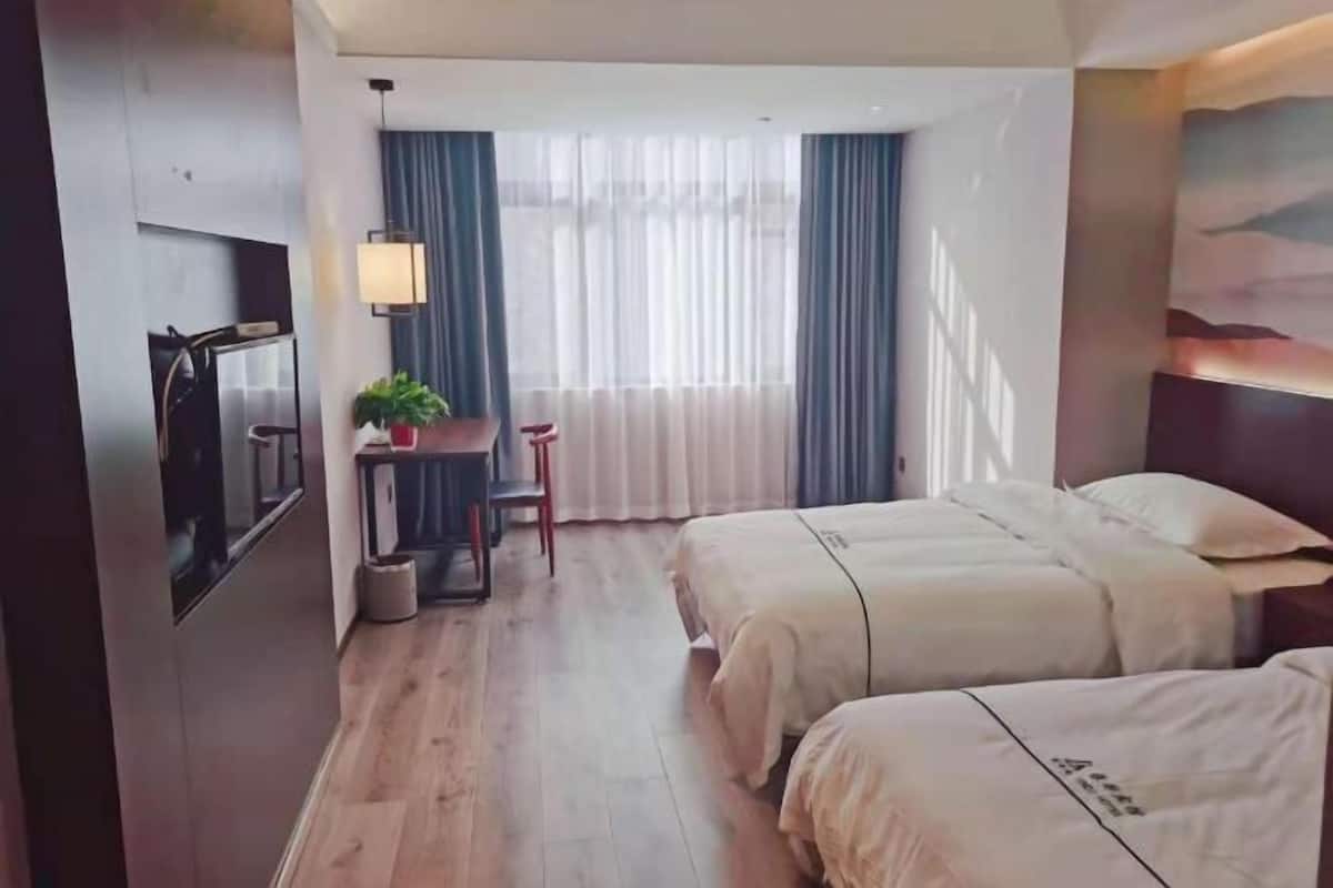 A hotel room with two beds, a desk, a chair, a lamp, and a large window with curtains.
