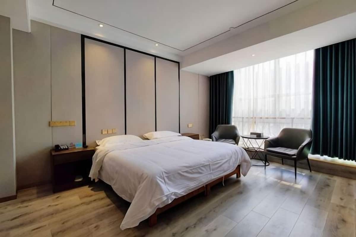 A modern hotel room with a large bed, a desk, a chair, and a view of the city through large windows.