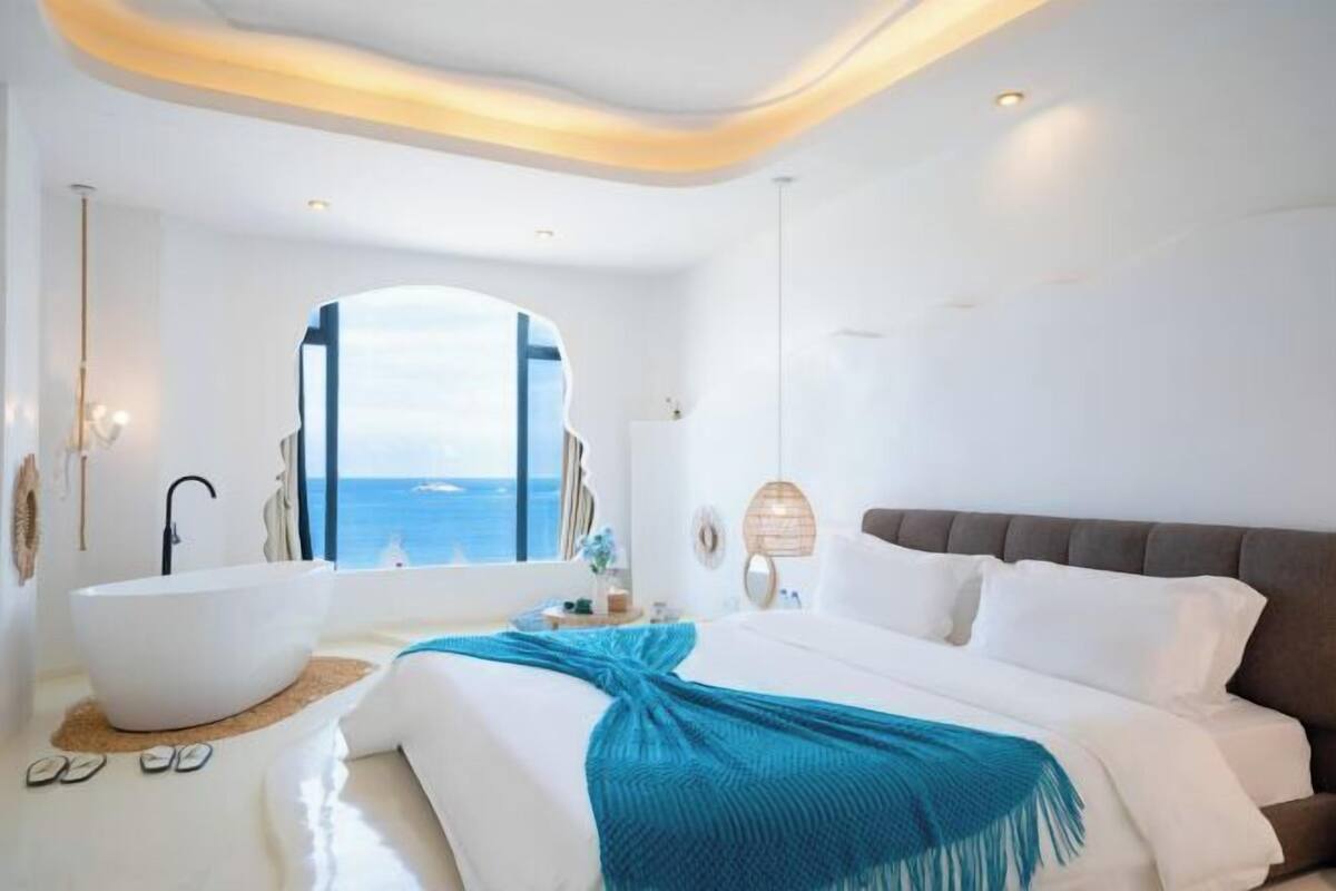 A modern hotel room with a large bed, a bathtub, and a view of the sea.