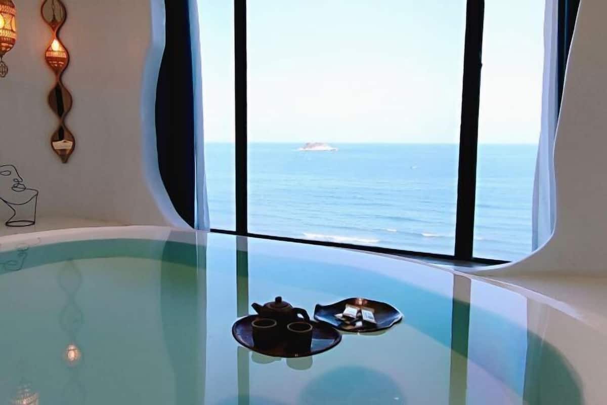 A glass-enclosed room with a view of the sea, a hot tub, and a table with tea set.