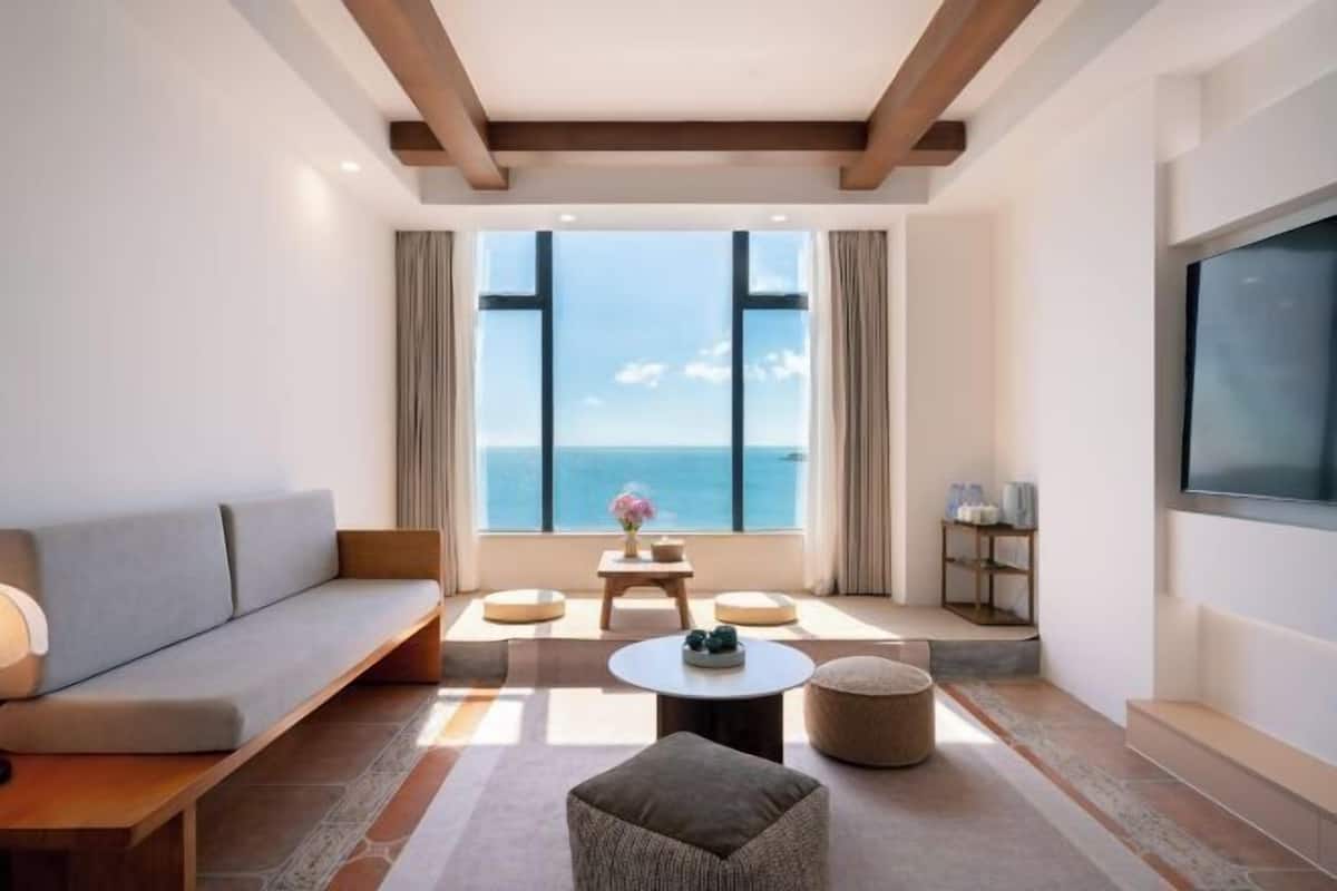 A modern hotel room with a large window, a sofa, a round coffee table, and a TV.