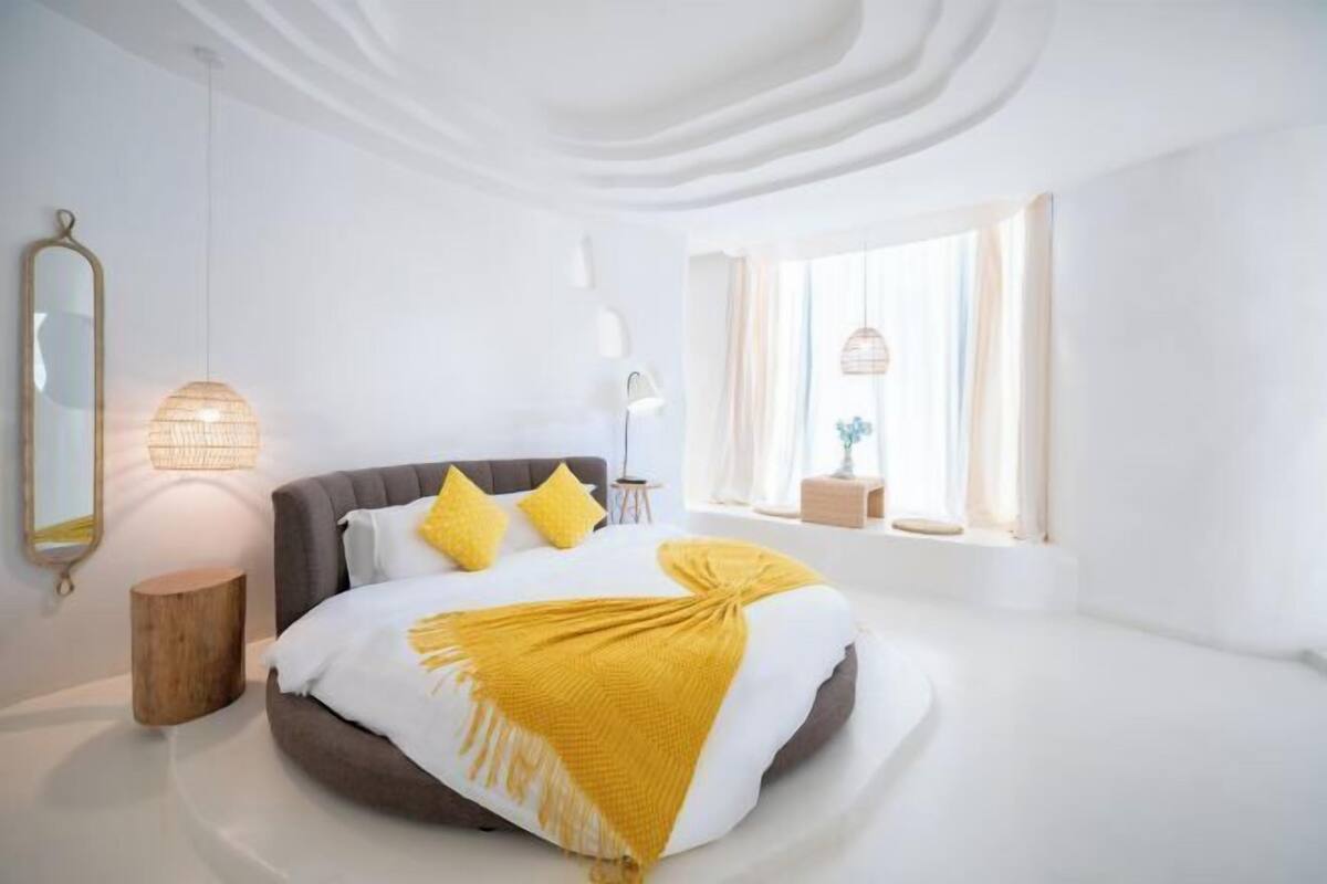 A modern bedroom with a round bed, a bedside lamp, a mirror, and a window with curtains.