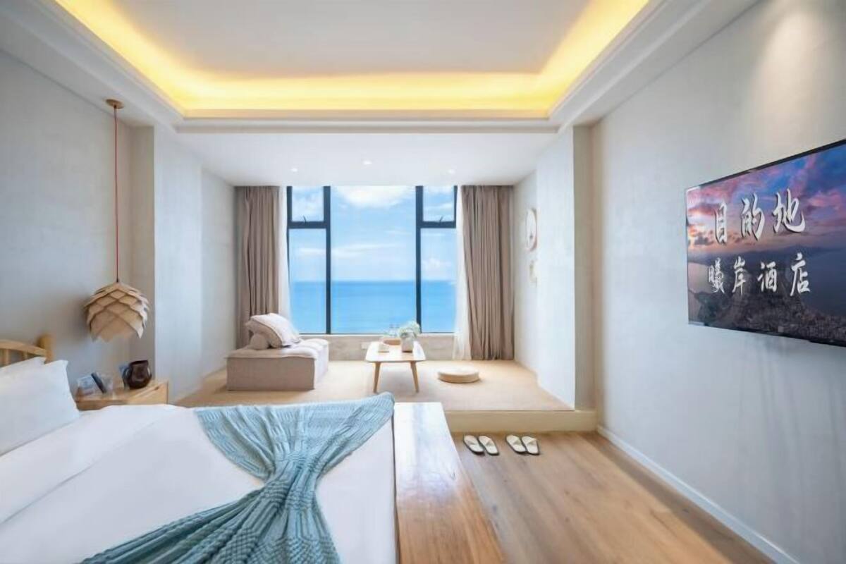 A modern hotel room with a large bed, a seating area with a view of the sea, and a wall-mounted TV.