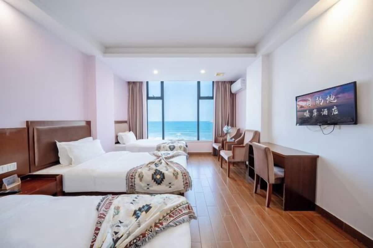 A hotel room with two beds, a desk, a chair, a TV, and a view of the sea.