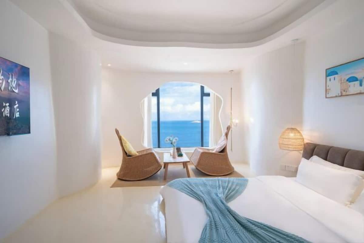 A modern hotel room with a large bed, two rocking chairs, a small table, and a view of the sea through a large window.