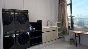 A compact laundry room with stacked washers and dryers, a sink, and a window with curtains. - Destination Xi An Hotel (Huidong)