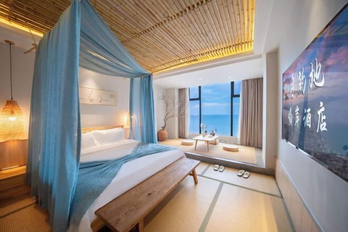 A modern hotel room with a large bed, a wooden bench, a view of the sea, and a large wall-mounted screen displaying text.