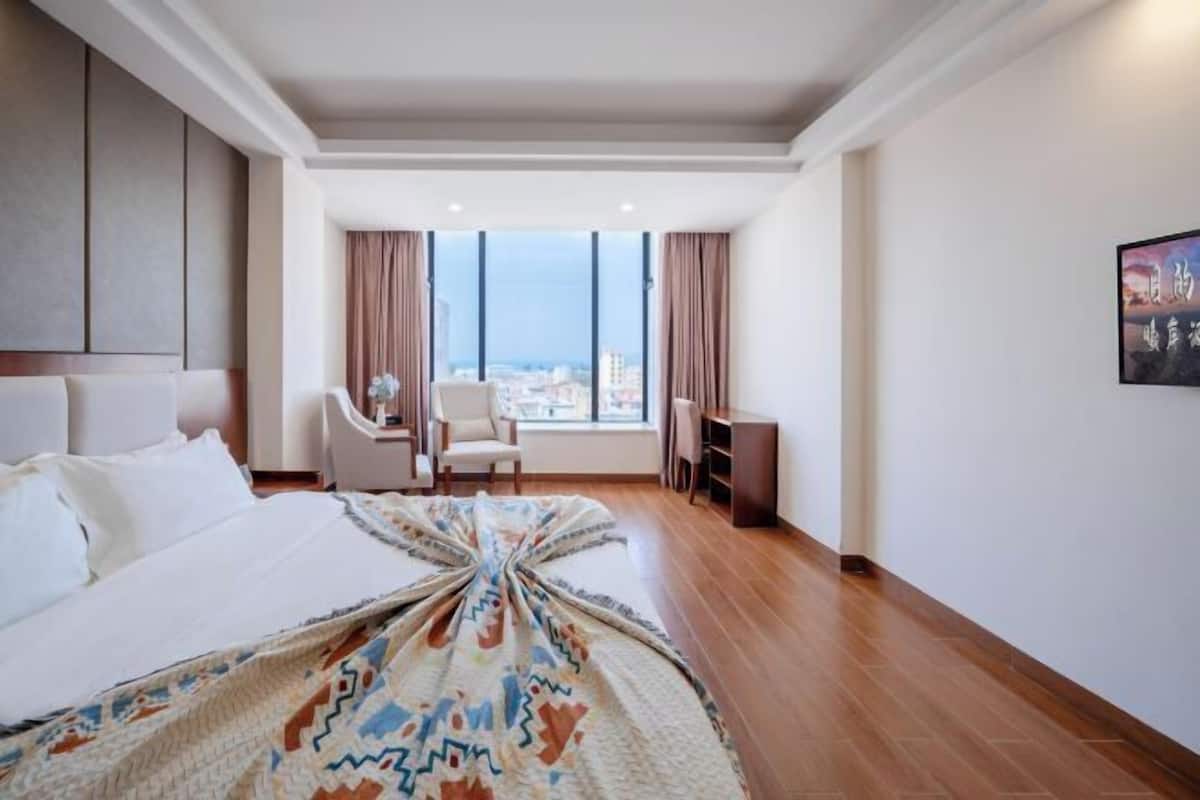 A hotel room with a large bed, a desk, a chair, and a view of the city through large windows.