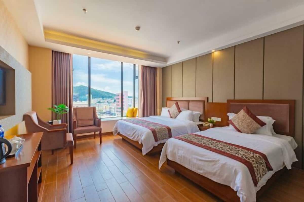 A hotel room with two beds, a desk, a chair, and a large window offering a city view.