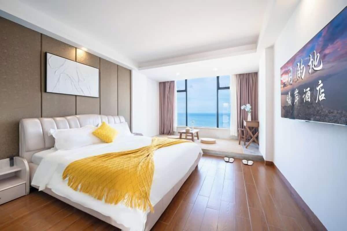 A modern hotel room with a large bed, a view of the sea, a seating area with a table and chairs, and a large artwork on the wall.