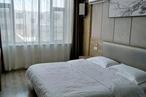 A hotel room with a bed, a window with sheer curtains, a headboard, and a wall-mounted air conditioning unit. - Jinhui Fashion Hotel (Baicheng)