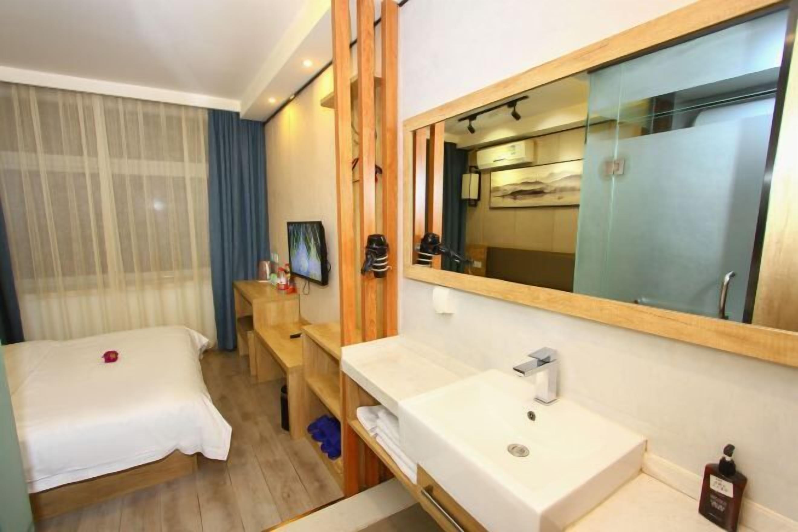 Habitación de hotel con cama, mesita de noche de madera, baño con lavabo y espejo, y ventana grande con cortinas.