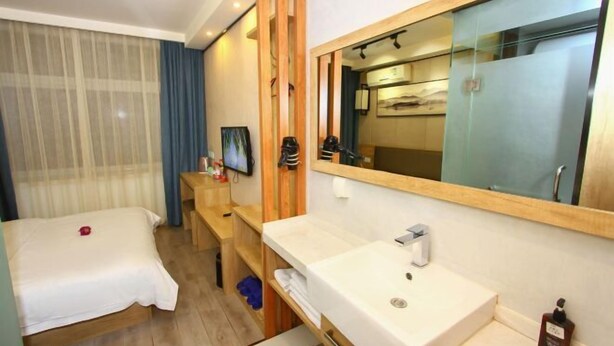 Habitación de hotel con cama, mesita de noche de madera, baño con lavabo y espejo, y ventana grande con cortinas.