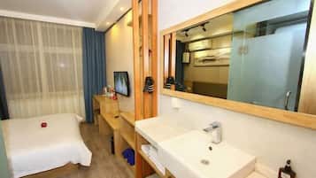 Habitación de hotel con cama, mesita de noche de madera, baño con lavabo y espejo, y ventana grande con cortinas.