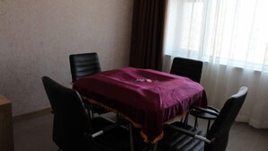 Una habitación con una mesa cubierta por un paño morado, cuatro sillas negras, una mesita de madera y una ventana con cortinas.