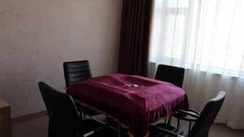 Una habitación con una mesa cubierta por un paño morado, cuatro sillas negras, una mesita de madera y una ventana con cortinas.