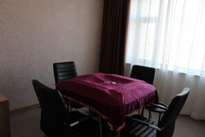 Una habitación con una mesa cubierta por un paño morado, cuatro sillas negras, una mesita de madera y una ventana con cortinas.