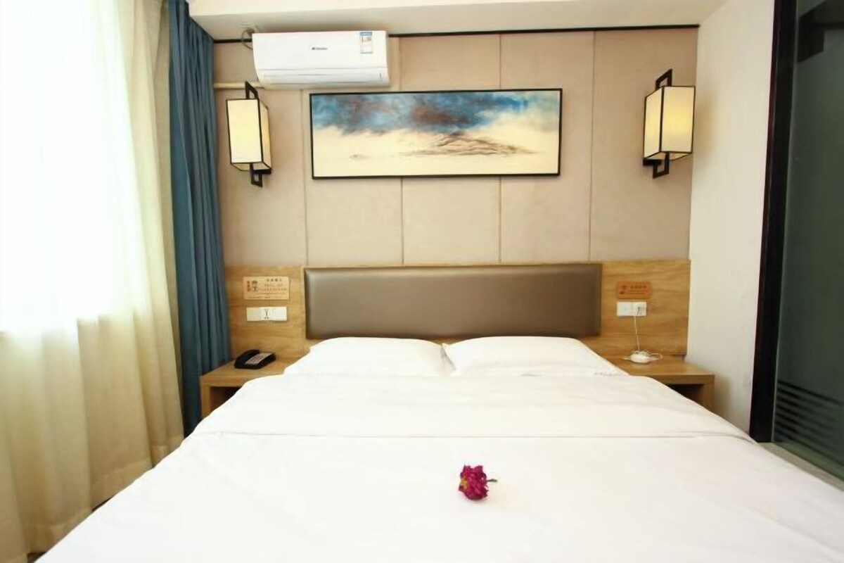 A hotel room with a bed, a nightstand with a phone, an air conditioner, and a painting on the wall.