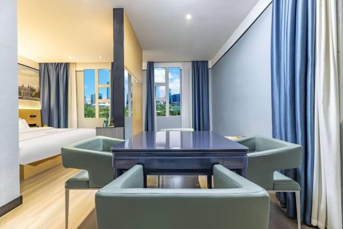 A modern hotel room with a large dining table, four chairs, a bed, and a view of a cityscape through the windows.