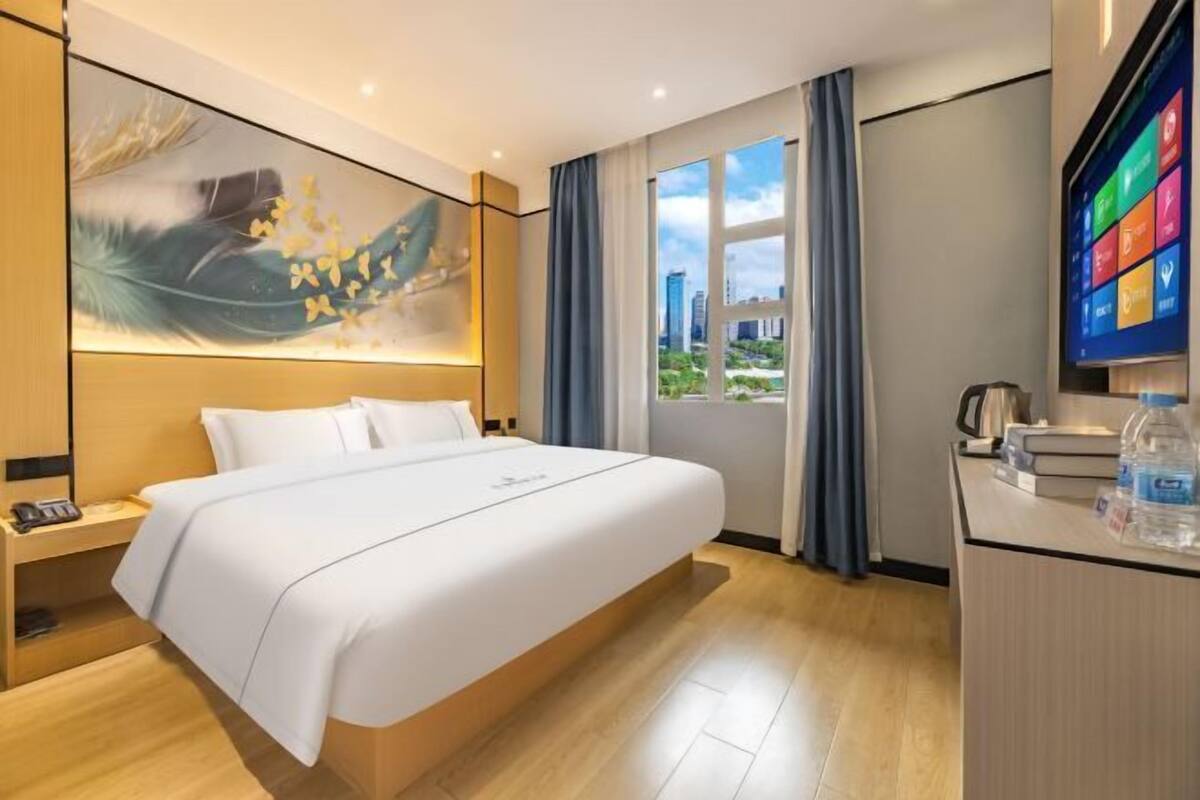 A hotel room with a large bed, a desk with a computer, a window with a city view, and a painting on the wall.