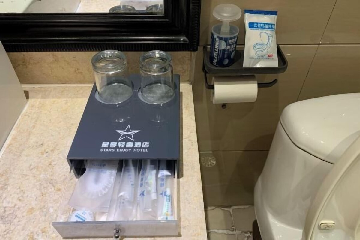 A Stars Enjoy Hotel restroom with a toilet paper holder, two glass containers, and a dispenser.