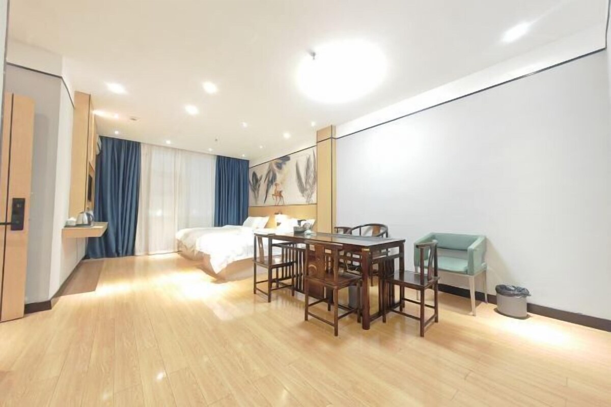 A modern hotel room with a wooden floor, a bed with white linens, a dining table with chairs, a small sofa, and a painting on the wall.