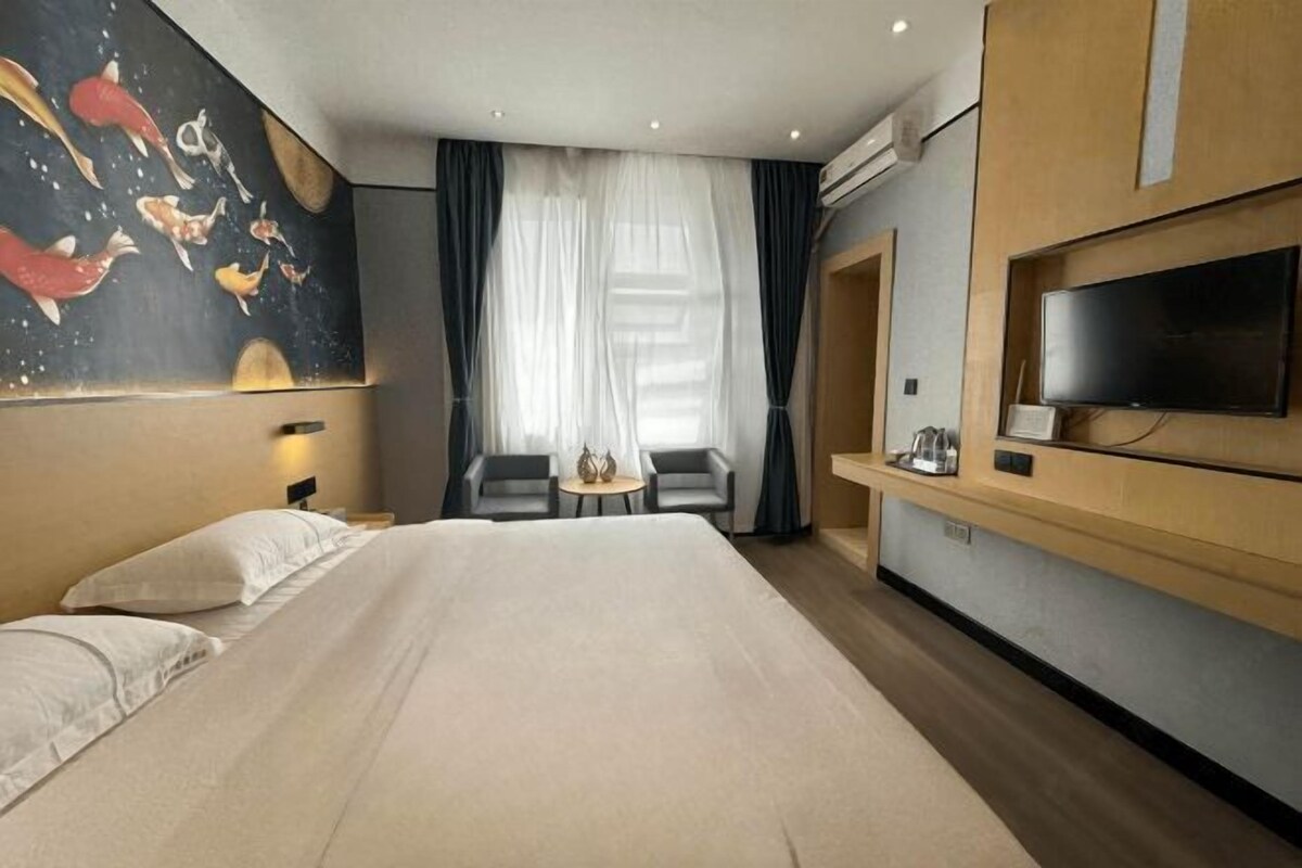 A hotel room with a large bed, a TV mounted on the wall, a small table with chairs, and a window with sheer curtains.