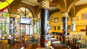 Restaurante com várias mesas e cadeiras, um pilar grande e uma televisão exibindo um evento esportivo.
