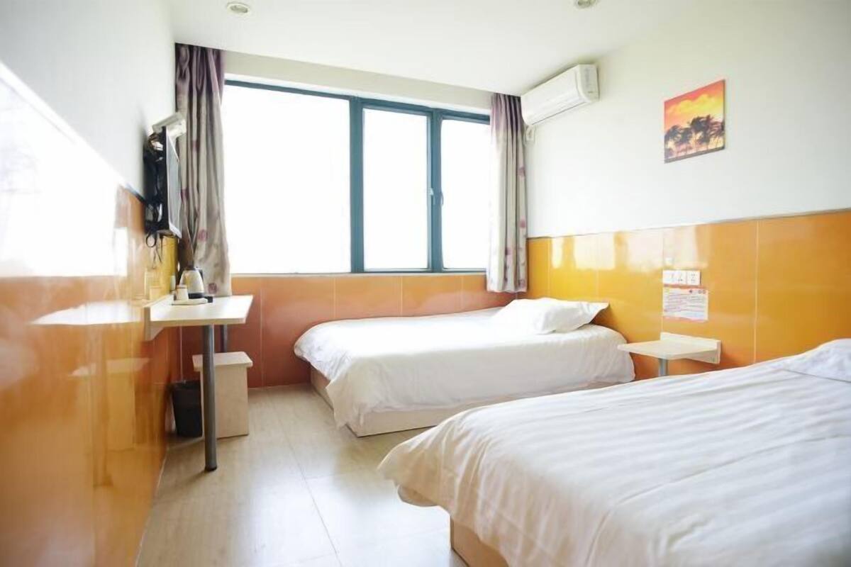 A hotel room with two single beds, a small table, a chair, a window with curtains, and a wall-mounted air conditioner.