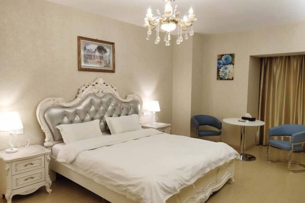 Habitación de hotel con una cama grande, una araña, un escritorio, dos sillas y obras de arte en las paredes.