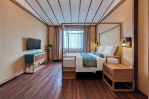 Una habitación de hotel moderna con piso de madera, una cama con estructura de madera, una mesita de noche, un televisor en la pared y una ventana con cortinas.