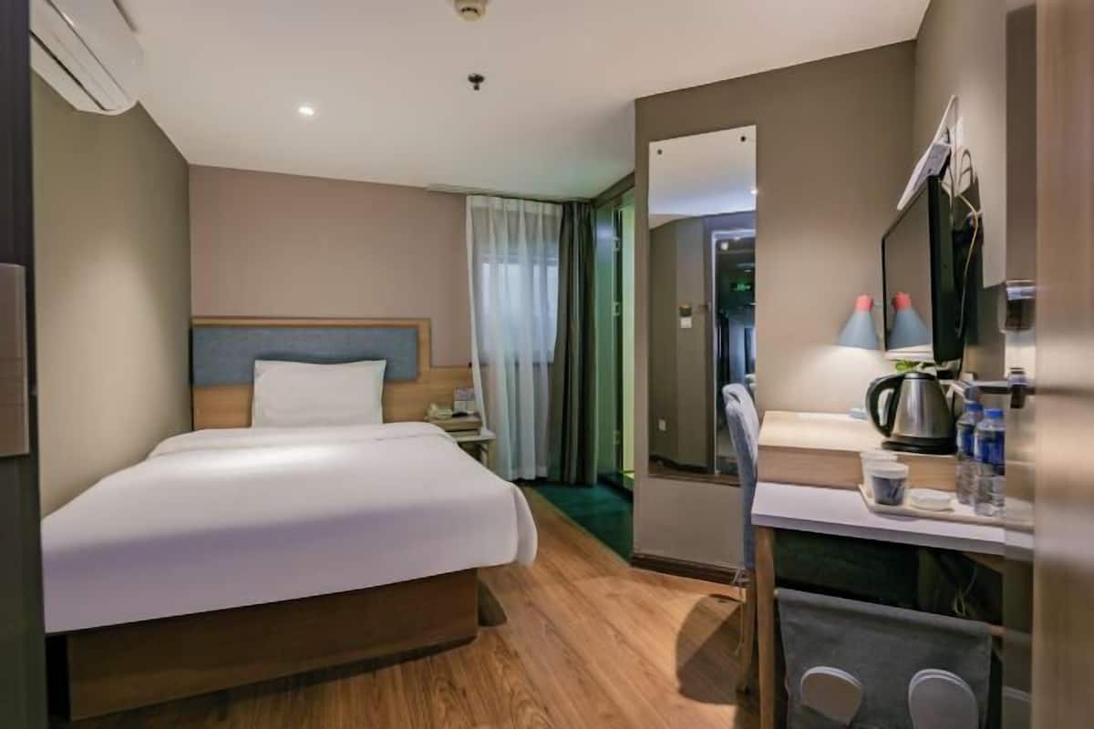 A hotel room with a bed, a desk with a chair, a mirror, a coffee maker, and a window with curtains.