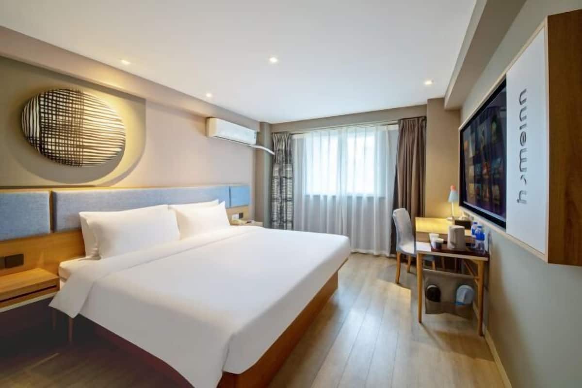 A hotel room with a large bed, a desk with a chair, a wall-mounted TV, and a window with curtains.
