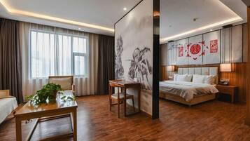 A hotel room with a wooden floor, a bed with white bedding, a desk with a plant, and a large painting on the wall.