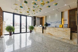 Zona de recepción de un hotel con suelo de mármol, mostrador de recepción, plantas en macetas y un ventanal grande.