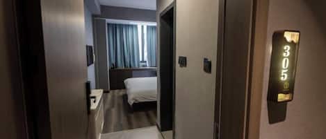 A hotel hallway with a bed, a desk, and a door with a number sign.