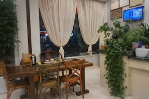 A dining area with a wooden table and chairs, a window with curtains, and a potted plant. - Sunshine Hostel (Nanping)
