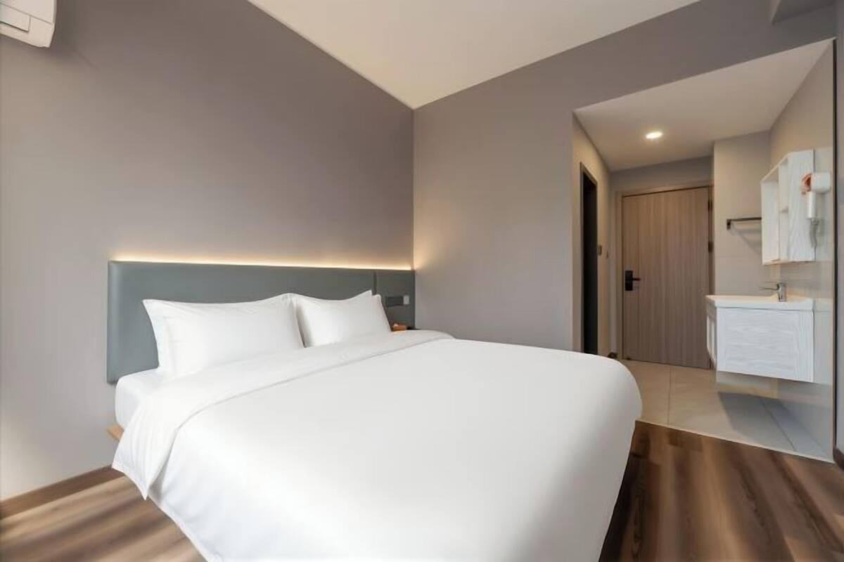 Une chambre d’hôtel moderne dotée d’un grand lit, d’une table de chevet, d’une tête de lit intégrée, d’un miroir et d’une salle de bain avec un lavabo et un porte-serviettes.