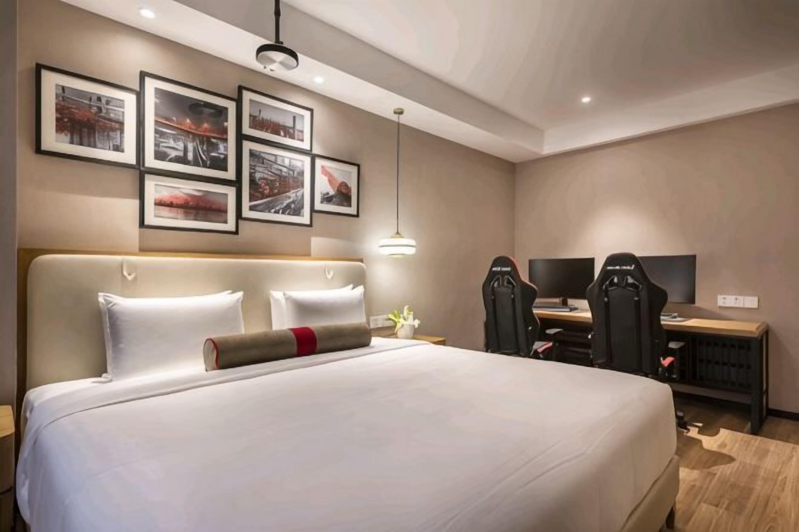 A hotel room with a large bed, a desk, and a chair. There are framed pictures on the wall and a lamp on the desk.