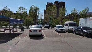 Ein Parkplatz mit mehreren Autos und einem weißen Lieferwagen. Im Hintergrund sind Bäume und ein Gebäude zu sehen.