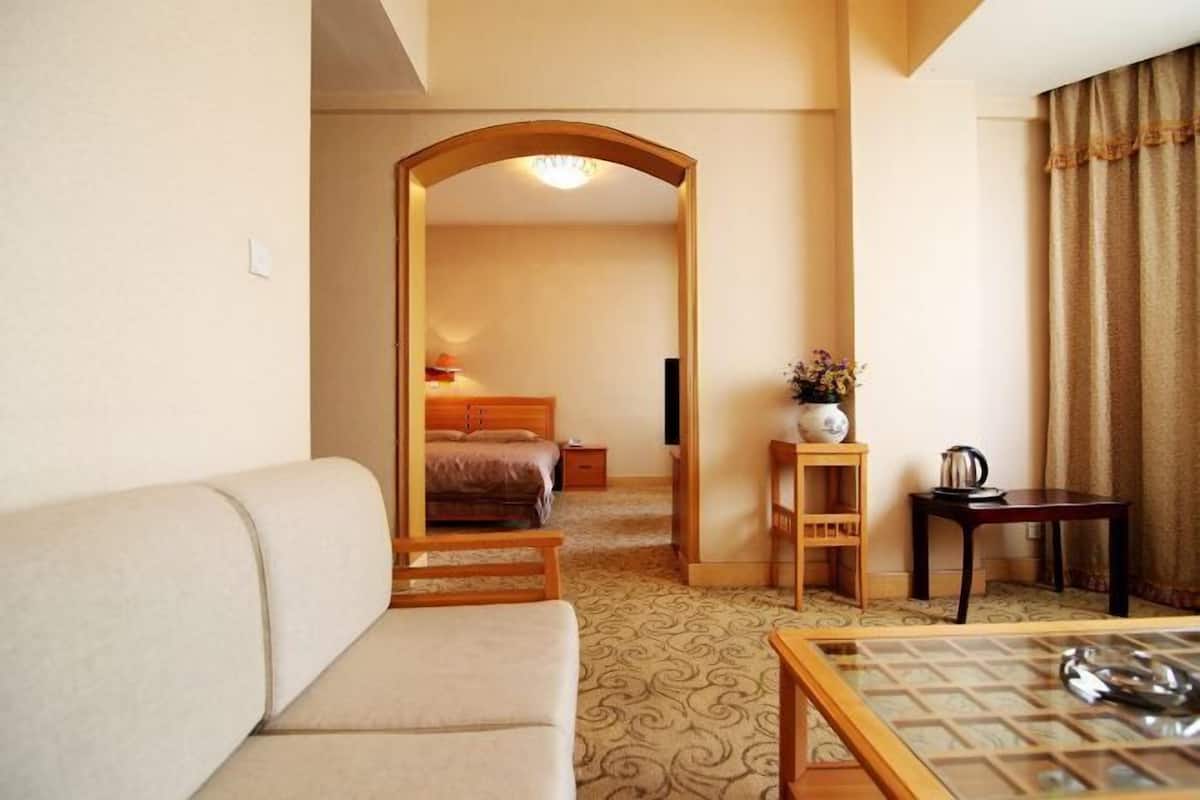 A hotel room with a sofa, a bed, a small table with a vase, a wooden chair, and a desk with a kettle.