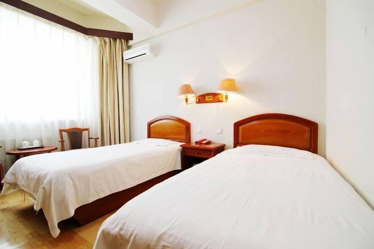 A hotel room with two single beds, a small table with a chair, a lamp, and a window with curtains.