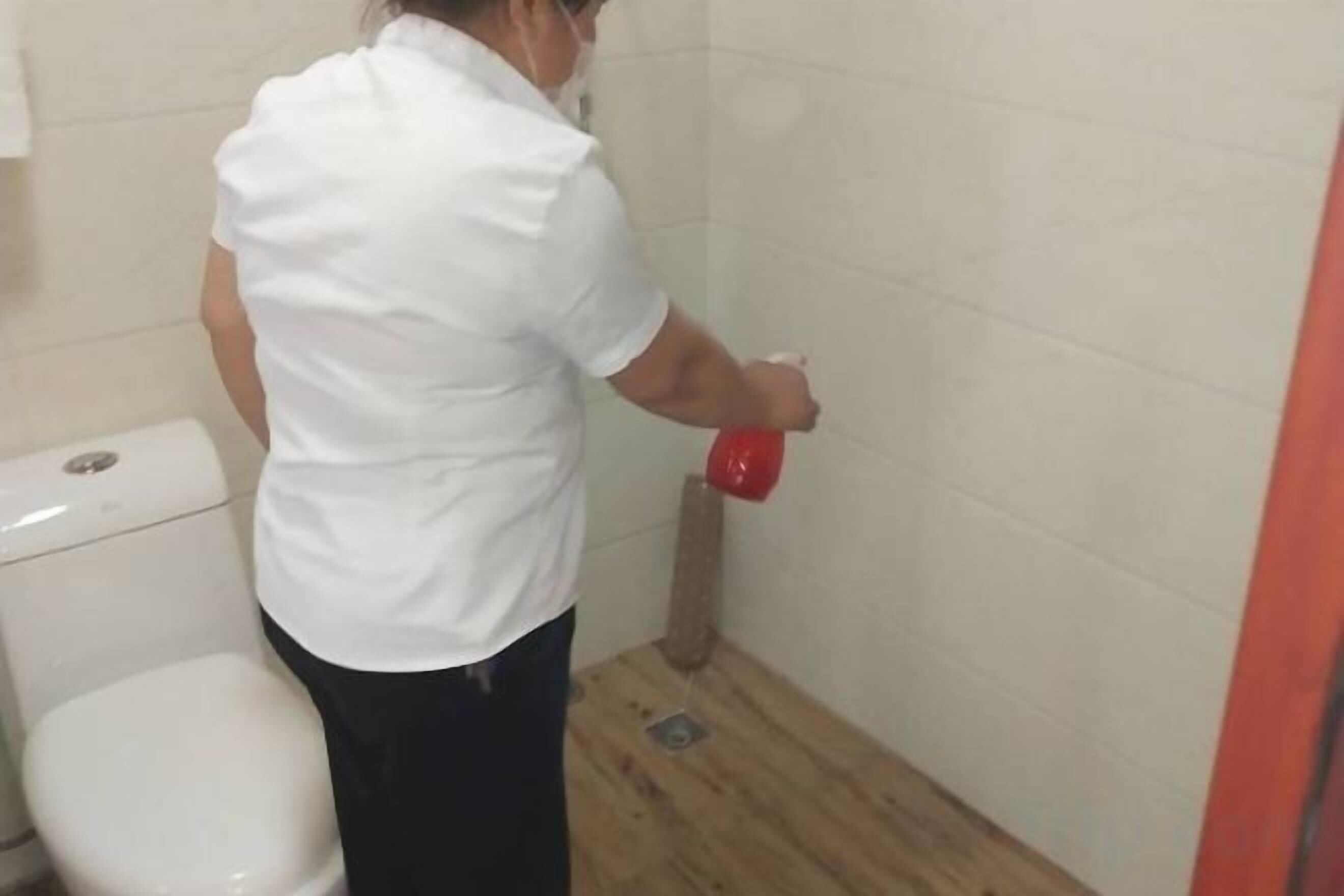 A person in a white shirt cleaning a bathroom with a red spray bottle.