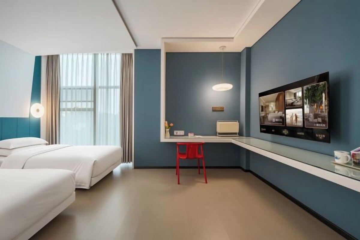 Habitación de hotel con dos camas, un escritorio con silla, un televisor montado en la pared y una ventana con cortinas.