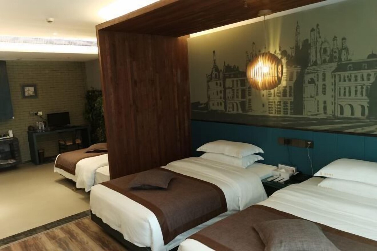 A hotel room with two beds, a wooden accent wall, a cityscape mural, and a desk with a lamp.