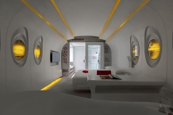 A modern, minimalist bedroom with a central bed, a bedside table, and a small TV. There are circular windows on the walls and a door leading to another room. - Mianyang 4D Future Hotel (Gaoshui Branch) (Mianyang)