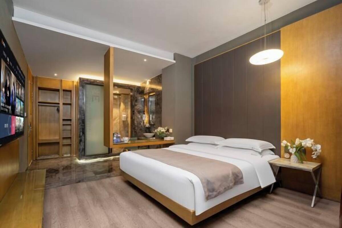 Ein modernes Hotelzimmer mit einem großen Bett, einem Schminktisch mit Spiegel, einem kleinen Tisch mit Blumen und einem Fernseher an der linken Wand.