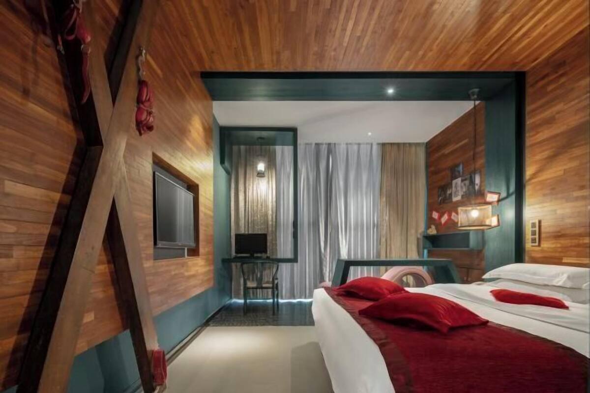 Ein modernes Hotelzimmer mit einer Wand in Holzoptik, einem Bett mit roter Bettwäsche, einem Schreibtisch mit Stuhl und Blick auf die Stadtlandschaft.